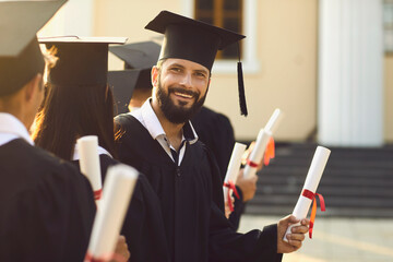 Portrait of happy male graduate holding his diploma standing in row with his groupmates near...