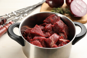Pot with raw meat, onion and metal skewers on white wooden table