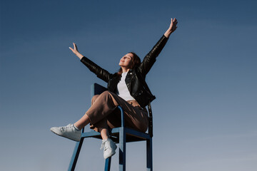 Happy young woman in black jacket sitting on chair with outstretched arms front of blue sky enjoying bright sunlight. Mental health, relaxation, freedom concept. Golden hour sunlight. Travel, airplane