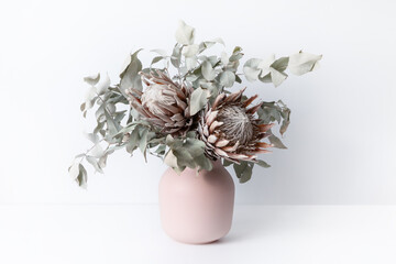 Beautiful dried flower arrangement in a stylish pink vase. In the flower bunch is pink King Proteas and Eucalyptus leaves, photographed on a white background.