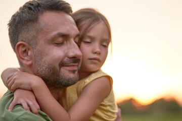 Close up shot of young father and adorable little daughter embracing each other, posing with eyes closed while spending time outdoors in the park on a summer day