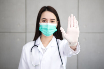 Fototapeta na wymiar Asian woman doctor who wears medical coat and face mask shows hand as “stop” sign in health protection concept at hospital. Stop epidemic of COVID-19.