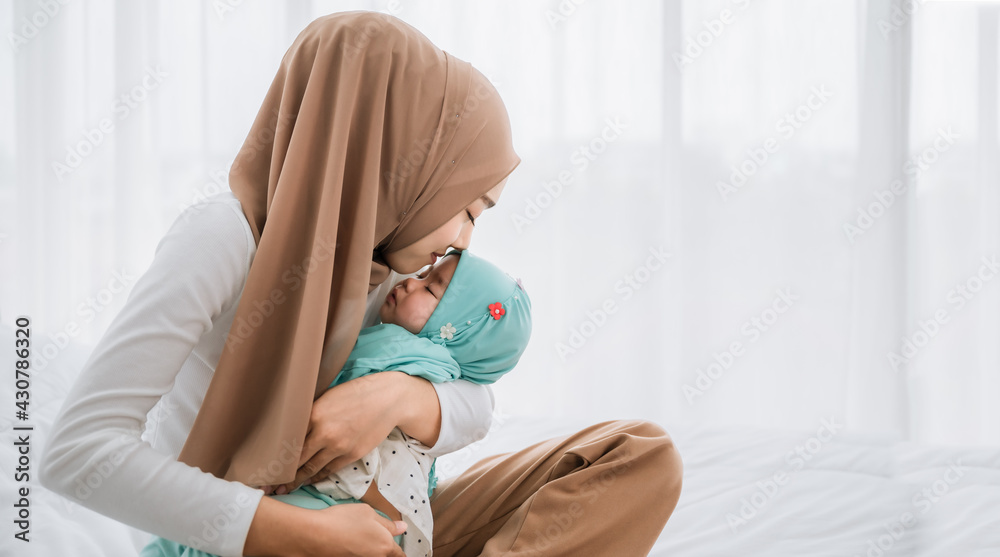 Wall mural happy asian muslim mother holding and kissing adorable little baby daughter wearing hijab in her arm