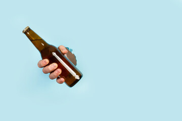 Mano de hombre sosteniendo una botella de cerveza a traves de un agujero en un papel celeste. Vista de frente. Copy space - obrazy, fototapety, plakaty