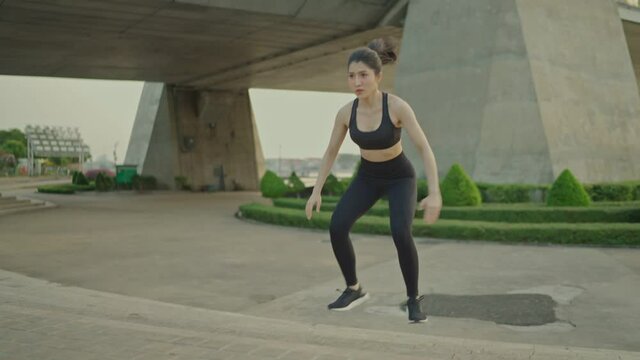 Slim Asian female athlete in black sportswear wearing exercise headphones jumps up stairs in a park near a bridge on a river, urban life.