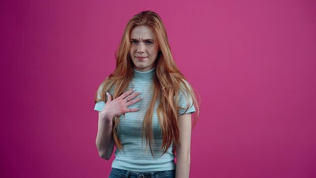 The portrait of a charismatic young woman who confuses her hand to her chest, I say, no, no, and she frowns, shows dislike. Freckled teenage girl in a blue T-shirt, isolated on a pink background. The