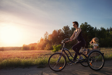 Fototapeta premium Young adult caucasian mom enjoy having leisure fun riding bicycle with cute adorable blond daughter holding wild field flower at scenic rural country road on bright sunny day. Countryside vacation