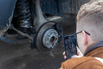 A man takes a video on his phone about the hydraulic brake of a passenger car. The brake disc is blurred, the man is focused