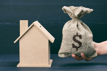 hand holds a canvas bag full of money and a wooden house on a blue background. The concept of...