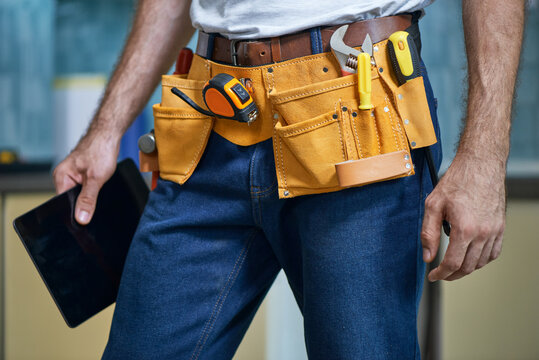 Cropped Shot Of Young Repairman Wearing A Tool Belt With Various Tools Holding Digital Tablet While Standing Indoors