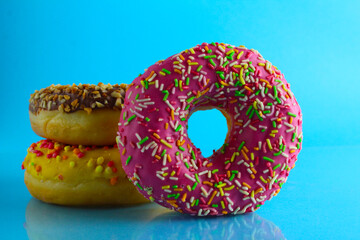 A still life eating sweet donut berliners in a pink glaze with a colored sprinkler stands next to a stack of doughnuts on a blue bright background with a place to text a picture for the table