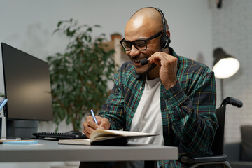 Disabled young man in wheelchair working at his working table with headset on