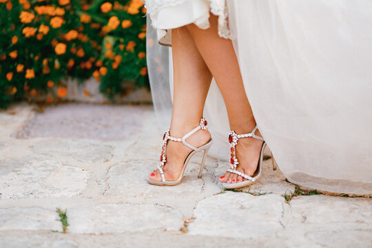 Bride's legs in stylish sandals with crystals with high heels peek out from under the wedding dress, close-up 