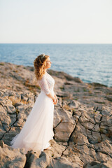 Fototapeta na wymiar A refined graceful bride with a diadem stands on a rocky seashore, the sun's rays fall on her face 