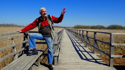 Fototapeta na wymiar sportlicher Senior posiert sitzend auf Holzsteg am Federsee vor Schilfgürtel unter blauem Himmel