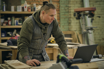 Concentrated adult craftsman checking information on laptop while working in carpentry workshop. profession, carpentry, technology and people concept
