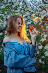 Young woman in denim jacket smiling while looking at an apple. The blonde hair glows with warm light. In the background a blooming garden. High quality photo