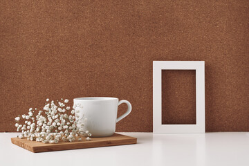 Frame, white flowers, cup, cork background. Mock up, copy space. Folk.