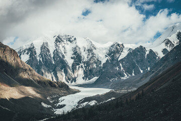 Beautiful mountain landscape with glacier tongue and great snowy mountains in sunlight in cloudy sky. Scenic alpine landscape with glacial tongue and high mountains with glaciers in low clouds.