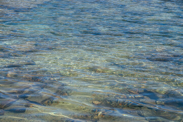 Meditative ripple of mountain lake. Beautiful relaxing background of blue green transparent water of glacial lake in sunlight. Sunny nature backdrop with many stones in clear water of glacier lake.