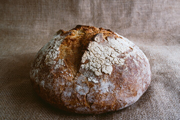 close up of fresh round loaf of healthy whole grain bread on brown jute burlap fabric background with white flour on top. top view of homemade bread on hessian fabric