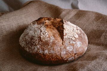 fresh round loaf of healthy whole grain bread on brown jute burlap fabric background with white flour on top. top view of homemade bread on hessian fabric