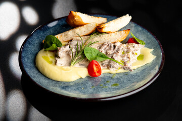 Meat in a creamy sauce with mashed potatoes and bread toast, painted with spinach leaves and cherry tomatoes, on a gray plate on a black background.