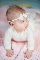 Fototapeta premium a cute baby in a fluffy dress looks away and stands on his handles. on the head of the banter and in white clothes. Blue eyes and fluffy hair