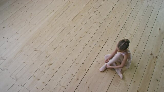 Top-view shot of pretty little ballerina sitting on wooden floor in empty ballet dance studio, tying pointe shoes preparing for classes