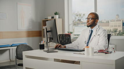 Experienced African American Male Doctor Wearing White Coat Working on Personal Computer at His Office. Medical Health Care Professional Working with Test Results, Patient Treatment Planning.