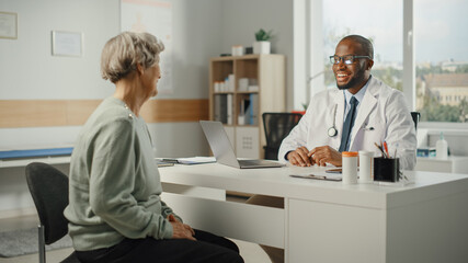 Obraz premium African American Family Doctor is Prescribing Medication to Senior Female Patient and Speaking with Her During Consultation in a Health Clinic. Physician Sitting Behind a Desk in Hospital Office.