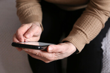 Elderly woman with smartphone sitting on sofa at home, mobile phone in female hands close up. Concept of online communication in retirement, sms, social media