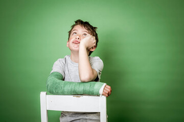 cool young schoolboy with broken arm and green plaster is sitting on white chair in front of green...