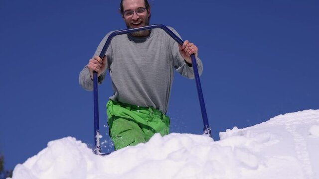 Close up shooting from below of a young man on the roof of a house in a bright sunny winter day shoveling down heaps of fresh snow towards camera.