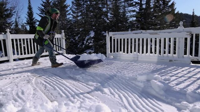 Snow removal in a sunny winter day. Slow motion shooting of a young man in fluo visibility jacket pushing around heaps of fresh snow from a veranda.
