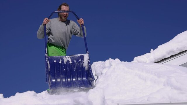 Wide angle view of a smiling young man on the roof of a house shoveling down heaps of fresh snow with a hand shovel. Beautiful, sunny winter day