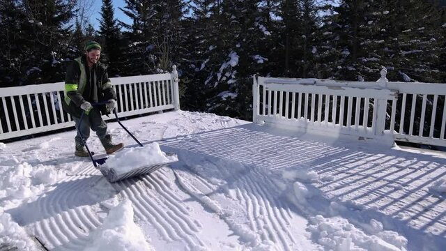 Snow removal in a sunny winter day. Strong young man in fluo visibility jacket pushing around heaps of fresh snow from a firts floor veranda.