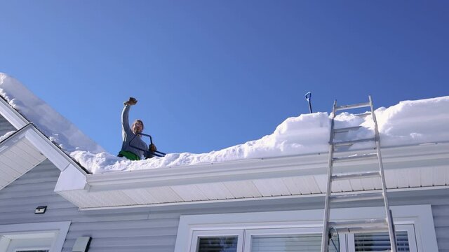 Slow motion shooting from below of a funny young man perched on the roof of a house removing fresh snow, throwing it down and exulting arm up.