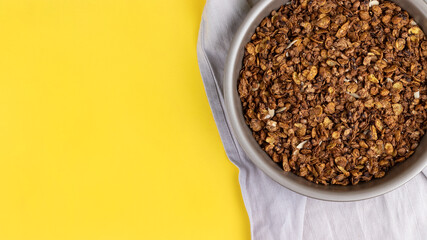 Organic homemade roasted granola cereal with oats, flax, almonds, chocolate in round baking tray with linen textile on bright yellow background. Top view, flat lay muesli wide banner with copy space