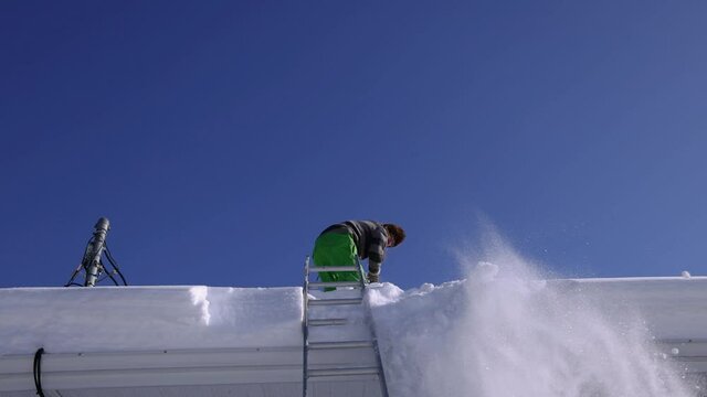 Slow motion shooting from below of a young man perched on the roof of a house removing fresh snow with a shovel and throwing it downwards.