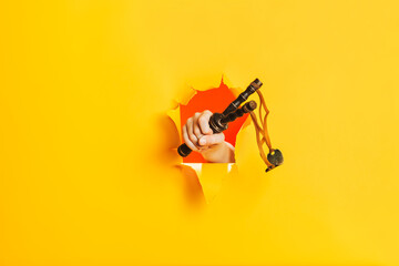 Naklejka premium Hand holds a wooden slingshot through a torn hole in a paper yellow wall. Shoot down concept. A side place for advertising.