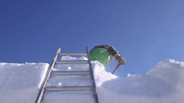 Slow motion shooting from below of a young man on the roof of a house removing fresh snow with a shovel and throwing it down towards camera.