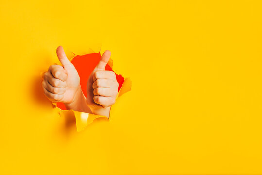 Two hands show OK gesture, approval, liking, acceptance through torn hole in yellow paper wall. Double thumbs up sign. A side place for text information, product or service advertising. Copy space.