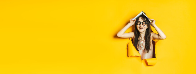 Little laughing nerd girl in black glasses and striped dress stands in the hole of torn colored yellow paper wall. The child holds an open book over his head. Space for text on the side.