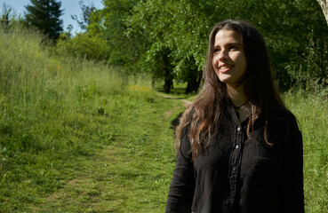 beautiful long-haired woman in black shirt, white pants and brown bag walking through nature with trees in the background and blue sky. teenager walking in the park watching the camera