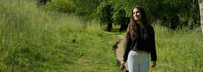 beautiful long-haired woman in black shirt, white pants and brown bag walking through nature with trees in the background and blue sky. teenager walking in the park watching the camera