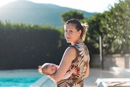 Happy Caucasian Mother With Imperfect Skin Holding Her Baby Girl In The Resort Garden Near The Swimming Pool - Family, Happiness, Real People And Love Concept