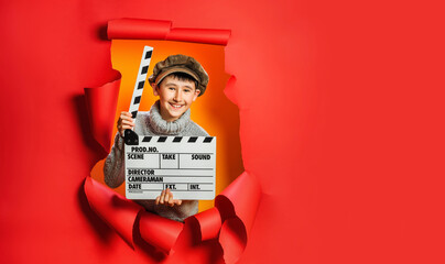 Little boy in knitted sweater and retro cap is playing movie maker holding wooden director's clapper board. Child is standing in hole in torn red paper wall. Side spot for your advertising, events.