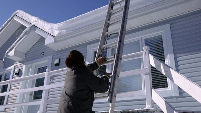 Long haired young man in fur hat carefully fixing and securing a professional extensible ladder to the roof of a house for a snow removal operation.