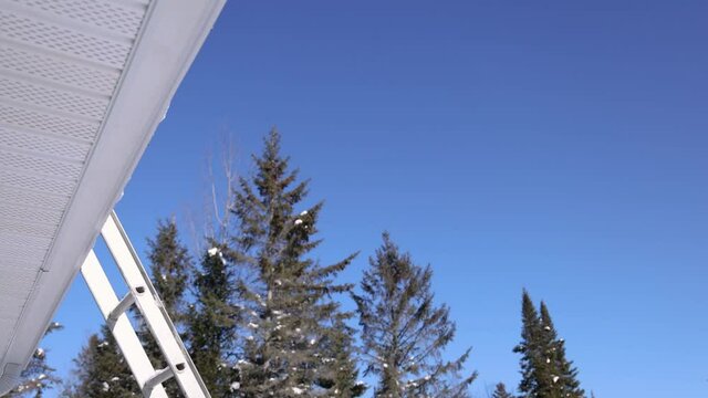 POV under the edge of a roof, pointing upwards. From the edge, heaps of snow come falling as someone on the roof shovels them down. Sunny winter day